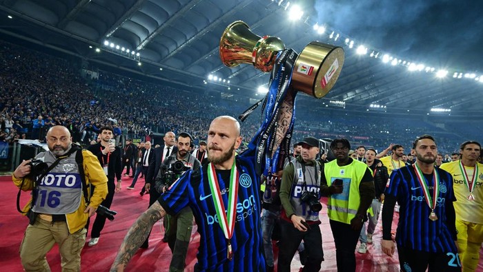 Footballer in blue-black striped jersey raises a large gold trophy above his head on a stadium pitch, photographers in the foreground and cheering crowd behind him.