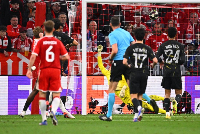Soccer scene: goalkeeper in yellow dives to stop ball at the near post as players in red and black react near the goal. Fans watch from the stands.