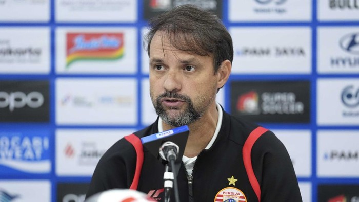 Man with dark hair and beard speaking at a press conference, wearing a black sports jacket with red stripes and a club crest, microphone in front of him with sponsor backdrop behind.
