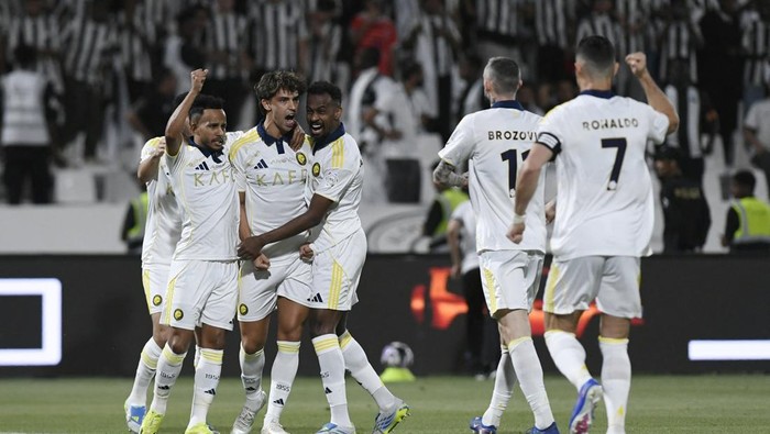 Soccer players in white uniforms celebrate a goal on the field, with number 7 visible on one back and a few teammates embracing in a group hug.