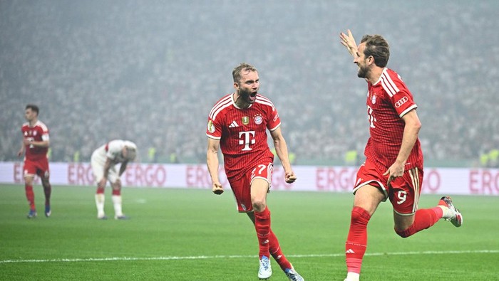 Two Bayern Munich players in red celebrate a goal on a rainy green pitch, with a gray crowd and advertising boards in the background.