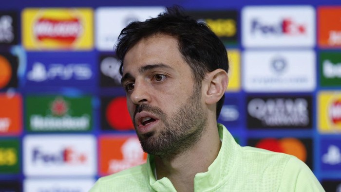 Male athlete speaking to reporters at a press conference with sponsor boards in the background, colorful logos visible behind him.