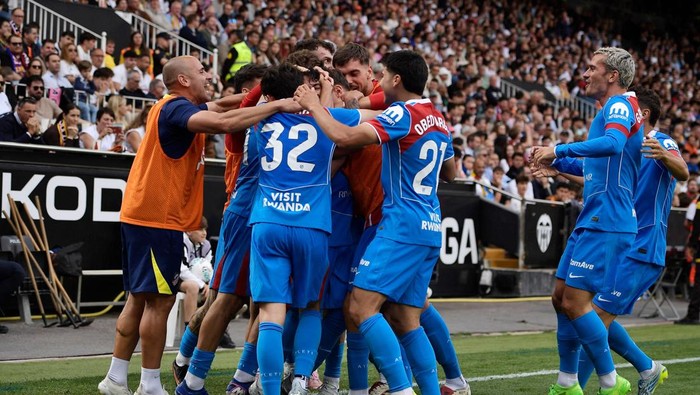 Soccer players in blue celebrate together on the field as fans cheer in the stands—group hug and high-fives after a play.