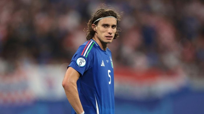 Soccer player in a blue jersey with the number 5 and a headband on the field during a match as fans watch from the stands.