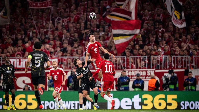 Red-clad Bayern player jumps to head the ball as opponents contest in a packed stadium.