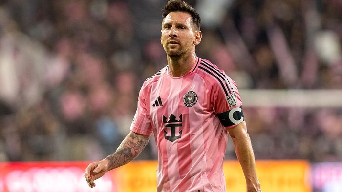 Footballer in a pink jersey with a captain's armband walking on a soccer field during a match.”] ,