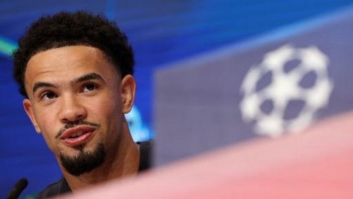 Close-up of a man speaking at a press conference, with a blurred UEFA Champions League logo in the background.
