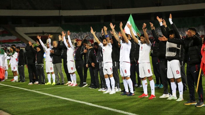 Soccer team in white jerseys standing in a line on the field with arms raised in unison.