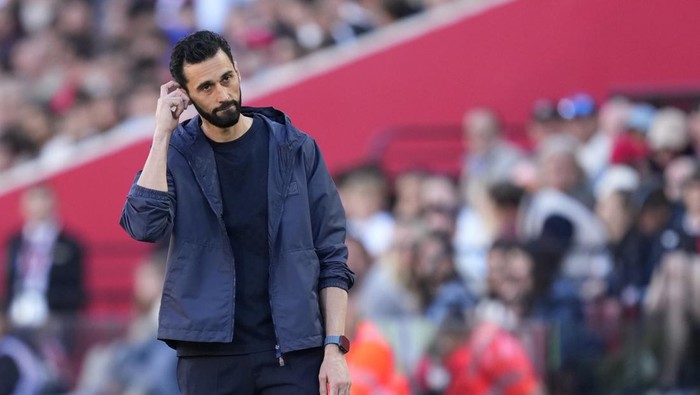 Bearded man in a blue jacket standing on a sports field, touching his ear as spectators watch from the blurred stands behind him.