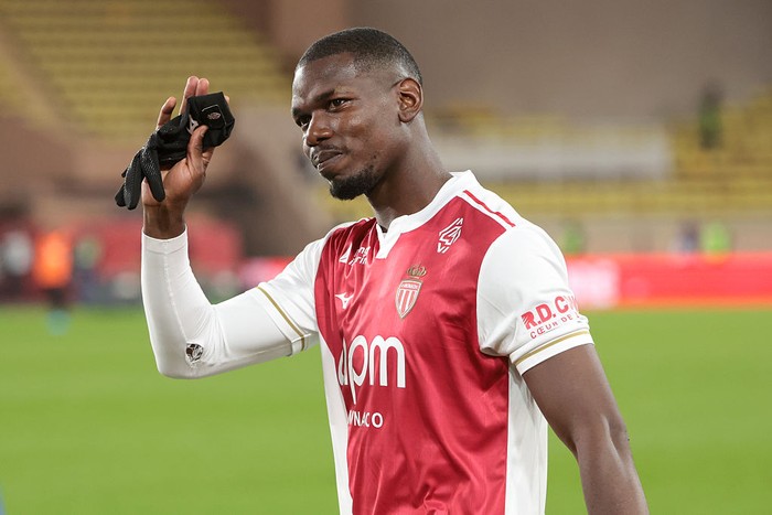 Soccer player in a red-and-white jersey lifts a small black camera toward his head on the field as fans watch in the background.
