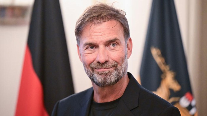 Portrait of a middle-aged man with gray beard in a dark blazer, standing in front of flags in a formal setting.
