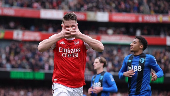Arsenal player in red kit covers his mouth with both hands on the pitch, with blue-jersey opponents nearby and a crowded stadium behind him.