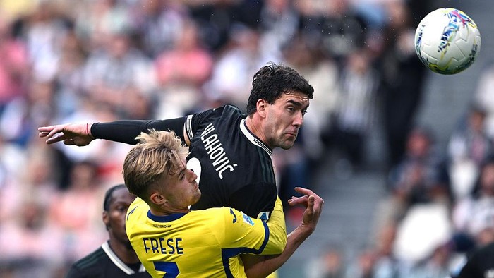 Two soccer players collide midair as they contest a high ball during a match; one in black, one in yellow boots nearby.