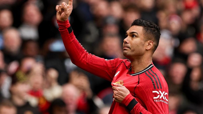 Soccer player in a red jersey points upward with one finger as a gesture of celebration, crowd blurred in the background