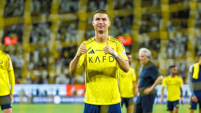 Soccer player in a yellow jersey on the field giving thumbs up, stadium crowd in the background.