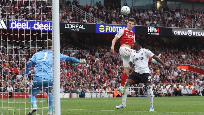 Soccer scene: Arsenal player in red jumps as the ball flies toward the goal, while a Fulham defender in white and black challenges; goalkeeper in blue ready at the left post.
