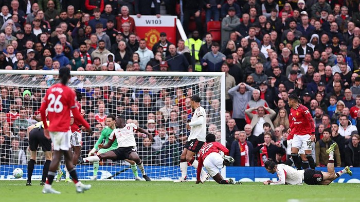 Soccer match near the goal as players in red contest a shot while the goalkeeper and defenders react, with a packed crowd behind the net.