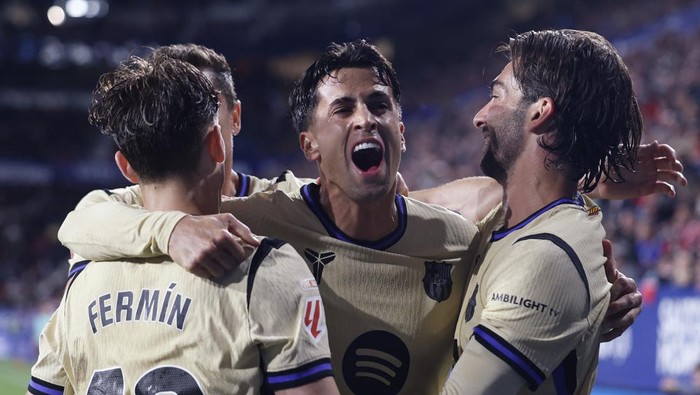 Three soccer teammates in beige jerseys celebrate with arms around each other on the field after a goal.