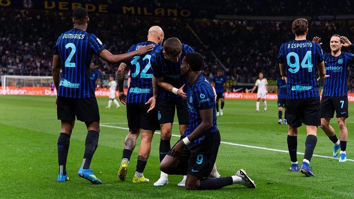 Inter Milan players celebrate a goal on the pitch, with one kneeling and teammates congratulating him.