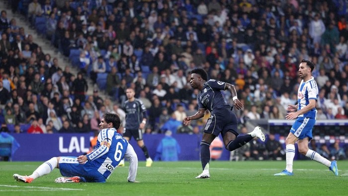 Soccer match moment: a blue-and-white team player slides on the grass while a dark-kit opponent runs toward the ball, with a packed crowd in the background.