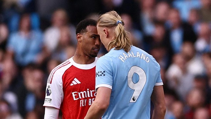 Close-up of two footballers from opposing teams staring each other on the pitch; Arsenal in red and Manchester City in light blue, Haaland wearing number 9 opposite them.