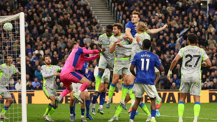 Soccer scene with players jumping around the goal as the goalkeeper in pink dives to reach the ball near the net while a crowded stadium looks on in the background.