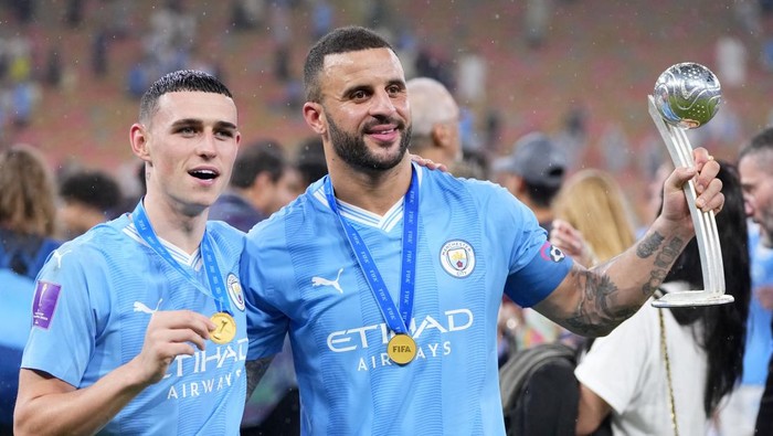 Two Manchester City players pose with medals, proudly displaying a large trophy during a celebration event in blue kits.