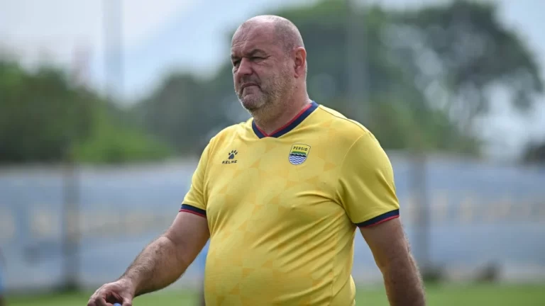 A bald man in a yellow soccer training jersey walking on a grassy practice field, looking ahead.