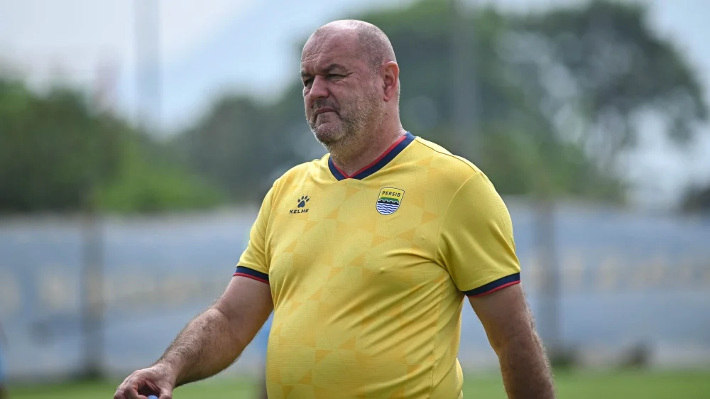 A bald man in a yellow soccer training jersey walking on a grassy practice field, looking ahead.