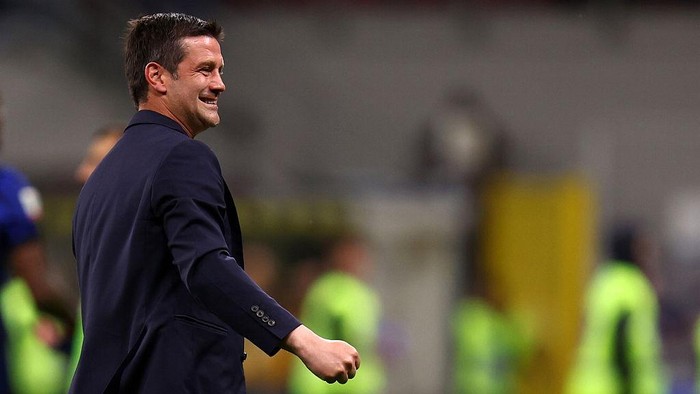 Man in a dark blazer walking across a sports field, smiling at the camera/arena.