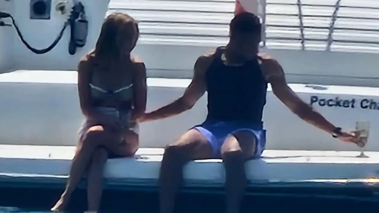 Two people sit on the edge of a white boat, facing away from the camera: a woman in a bikini and a man in a black sleeveless shirt with blue shorts.