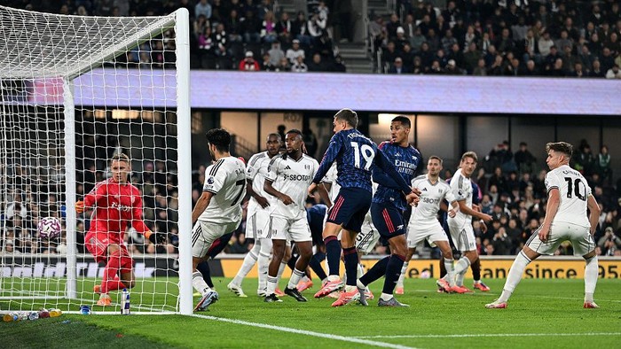 Soccer match near the goal: white-clad players defend as a blue attacker presses toward the net, goalkeeper in red watching from the left post.