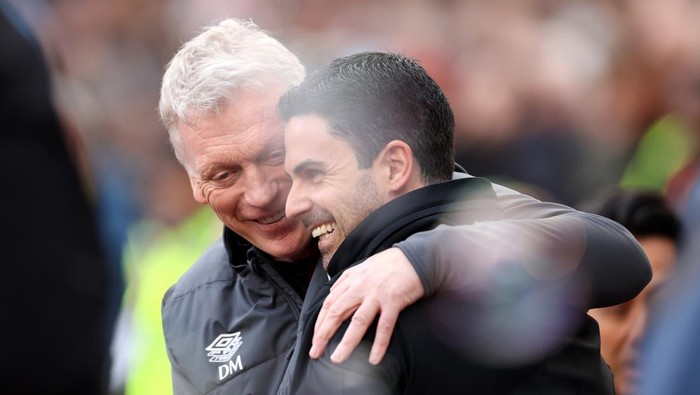Two men in sports jackets share a warm hug at a crowded sports event.