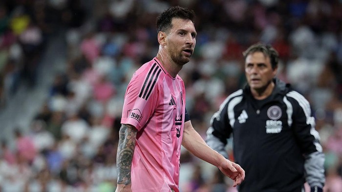Soccer player in a pink jersey walking on the field with a coach in a black tracksuit in the background.