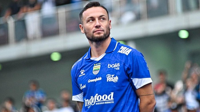 Male soccer player in a blue jersey standing on a field, looking to the right, with a blurred stadium background.
