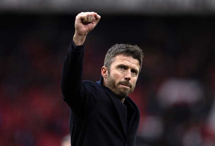 Man in a dark coat raises a clenched fist in the air on a stadium field, expression focused and victorious.