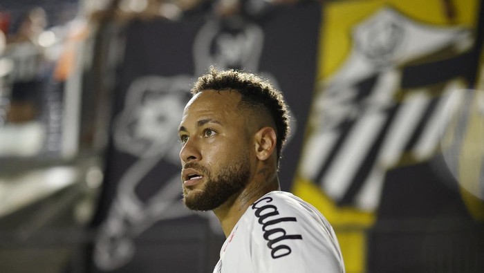 Close-up of a male soccer player with a beard in a white jersey looking to the left, stadium backdrop blurred behind him.