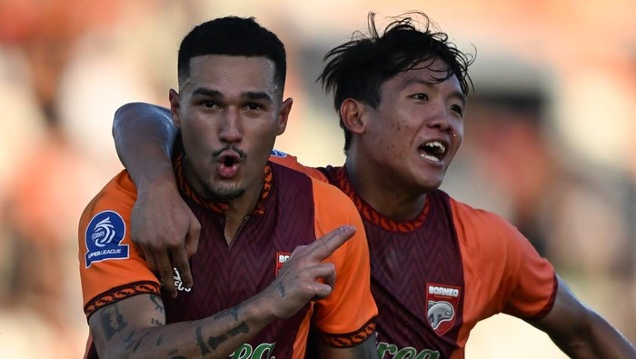 Two football players in maroon-and-orange kits celebrate a goal with arms around each other; one is shouting and pointing.