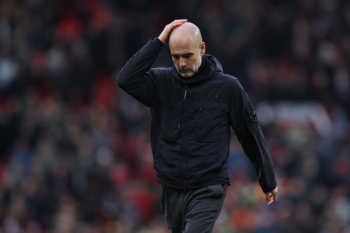 Soccer manager with hand on his head, walking off a pitch in a dark jacket.