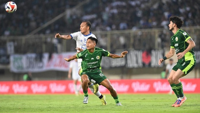 Soccer players in green and white compete for a ball during a match on a grassy field; ball is in the air to the left.