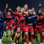 A soccer team in red and black uniforms celebrates on the field, arms raised and smiling after a win.
