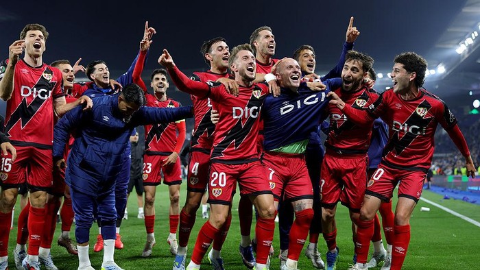 A soccer team in red and black uniforms celebrates on the field, arms raised and smiling after a win.