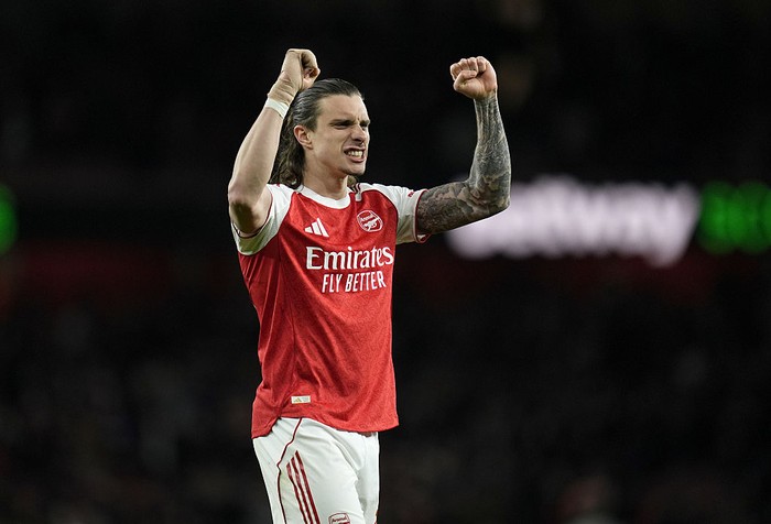Arsenal footballer in a red and white kit raising both arms in celebration on the pitch.