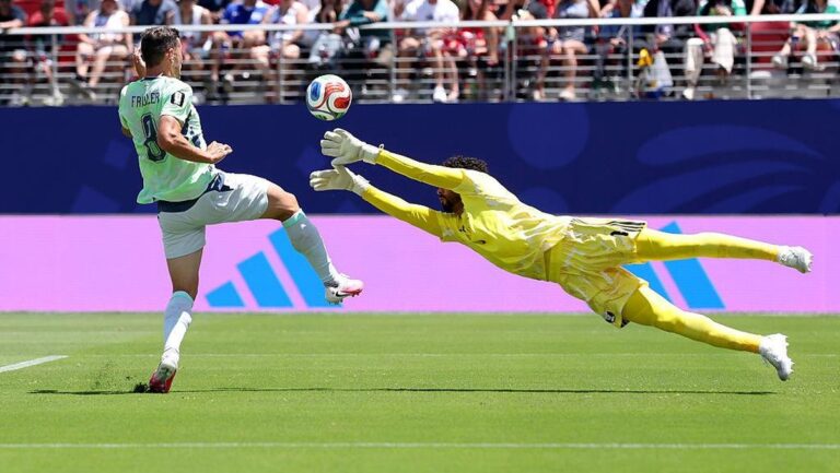 Soccer goalkeeper in yellow dives horizontally to block a ball as an opposing player in a light green kit attacks toward goal, with spectators in the stands behind.