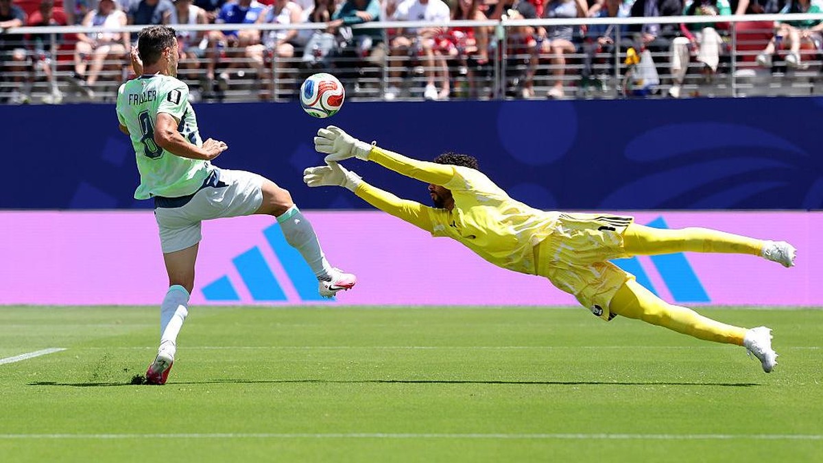 Soccer goalkeeper in yellow dives horizontally to block a ball as an opposing player in a light green kit attacks toward goal, with spectators in the stands behind.