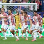 Croatian men's national soccer team players celebrate running across the field after a goal, white shorts, red‑white checker shirts, goal net in background.