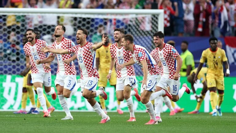 Croatian men's national soccer team players celebrate running across the field after a goal, white shorts, red‑white checker shirts, goal net in background.