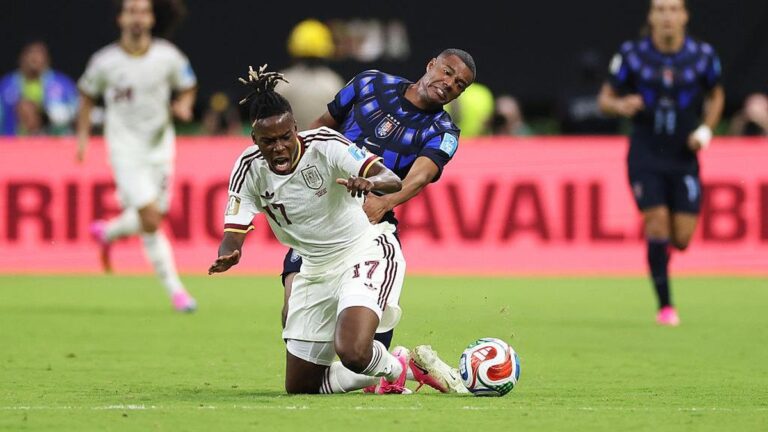 Soccer player in white kit (number 17) slides for the ball as a blue-clad opponent tackles from behind on a green field.