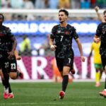 Group of football players in black kits celebrate a goal on the pitch, numbers 22, 23, 7 and 4 visible in front.
