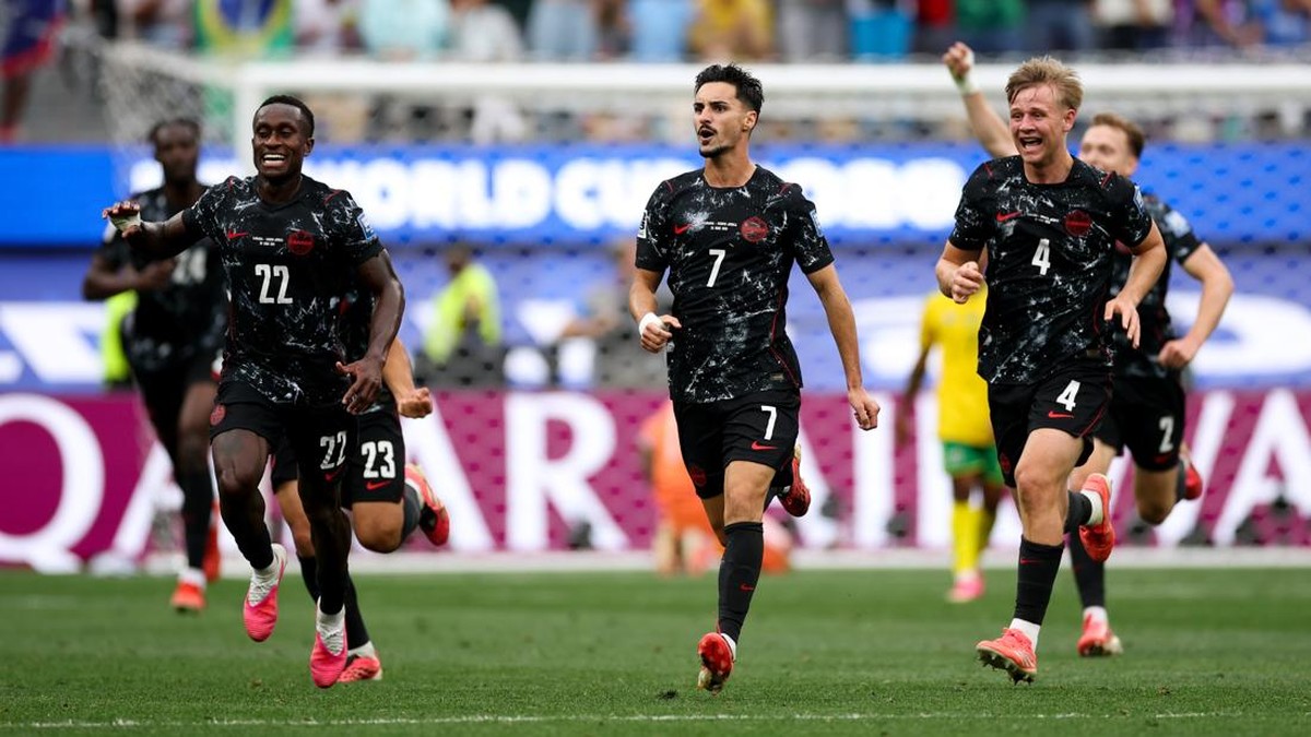 Group of football players in black kits celebrate a goal on the pitch, numbers 22, 23, 7 and 4 visible in front.